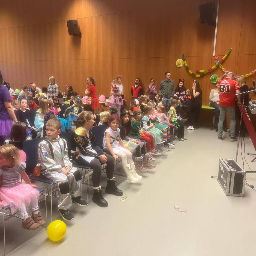 Eine Gruppe von Kindern und Erwachsenen in festlichen Kostümen sitzt in einem Raum, während ein Mann in einem roten Shirt neben einem Mikrofon steht. Ein gelber Ballon liegt auf dem Boden, und Lautsprecher sind an der Wand montiert.