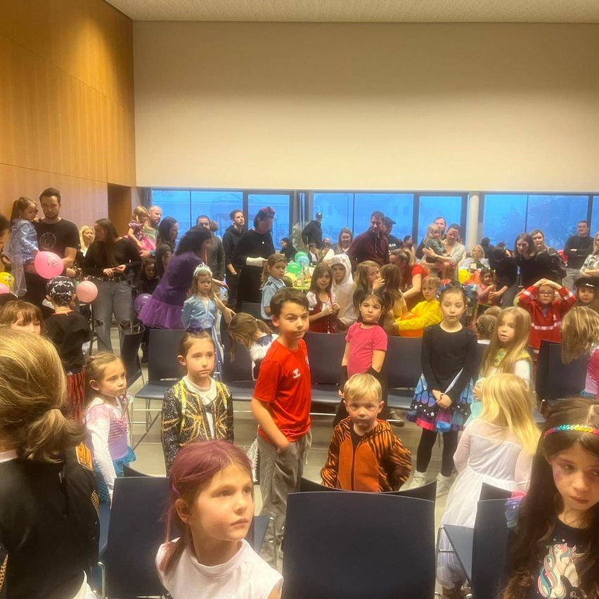 Eine Gruppe von Kindern und Erwachsenen versammelt sich in einem Raum, einige in Kostümen und mit Luftballons. Die Kinder sitzen in Stühlen, andere stehen herum. Der Raum hat Holzwände und große Fenster mit Blick auf Bäume draußen.
