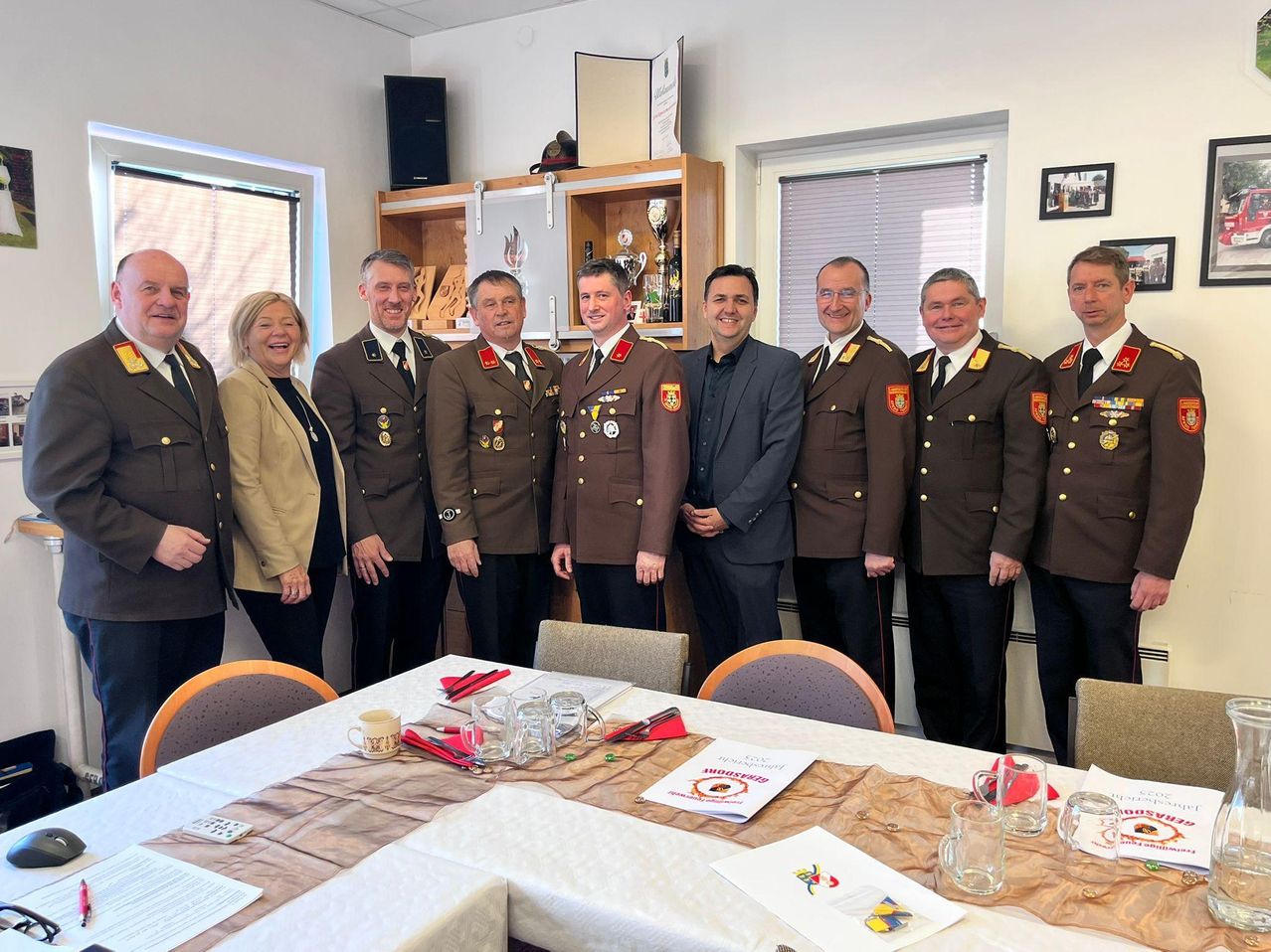 Eine Gruppe von Militäroffizieren und einer Frau steht lächelnd in einem Raum. Sie stehen vor einem Tisch mit Papieren und einem Pokal.