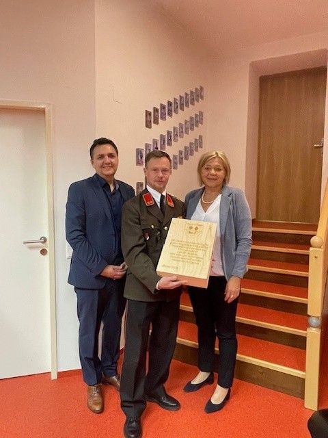 Drei Personen stehen in einem Raum im Innenbereich und lächeln. Ein Mann in Uniform hält eine Plakette, während zwei andere neben ihm stehen. Hinter ihnen zeigt eine Wand verschiedene Bilder, und eine Treppe führt zu einer Holztür.