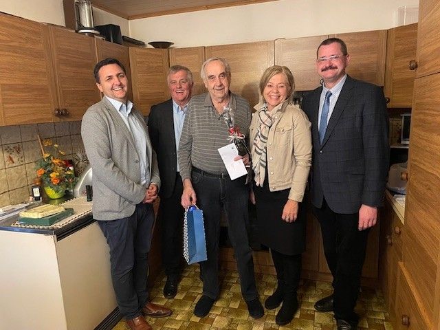 Fünf Menschen stehen in einer Küche, lächeln und posieren für ein Foto. Ein Mann hält eine Flasche und Papier, ein anderer eine Blumenbouquet, und eine Frau trägt einen Schal.