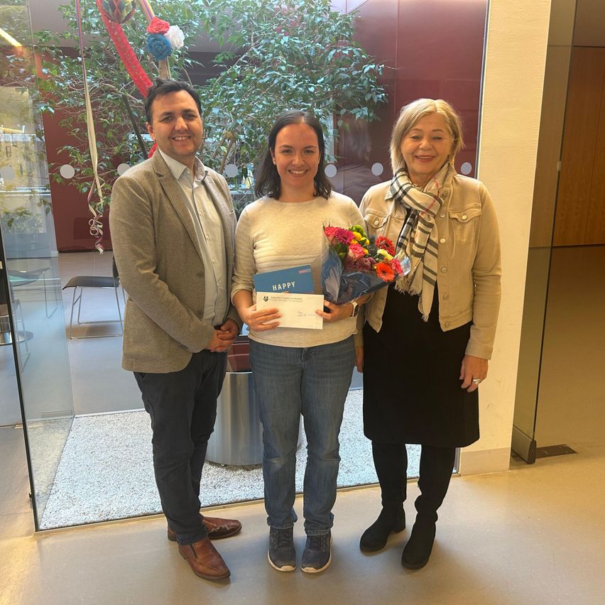 Drei Personen stehen lächelnd drinnen zusammen. Die Frau in der Mitte hält ein Zertifikat und einen Blumenstrauß. Der Mann links trägt einen Anzug und die Frau rechts einen Schal. Sie stehen neben einer Glaswand mit einer Pflanze und Dekorationen.