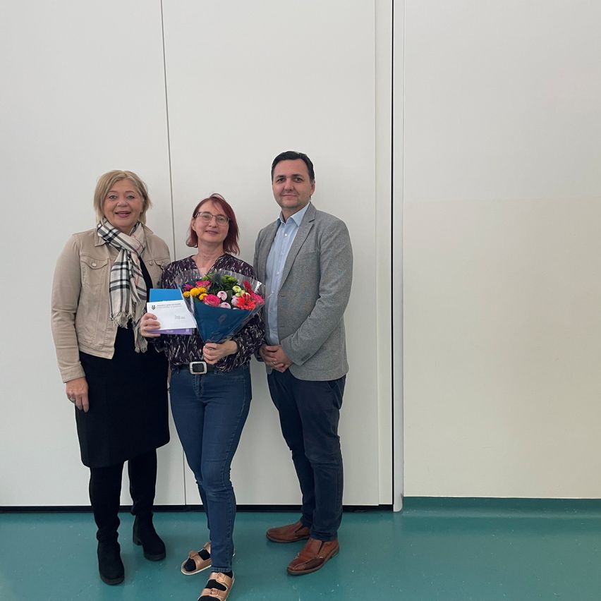 Drei Personen, zwei Frauen und ein Mann, stehen lächelnd vor einer weißen Wand. Die Frau in der Mitte hält einen Blumenstrauß.