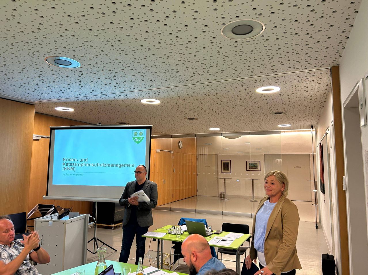 Ein Mann präsentiert ein Dokument bei einer Besprechung, mit einer Projektionsleinwand hinter ihm. Es stehen zwei Frauen und ein Mann sitzt an einem grünen Tisch mit einem Laptop.