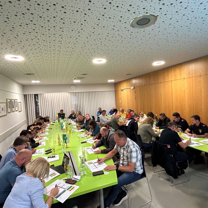 In einem großen Sitzungssaal sitzen mehrere Personen an grünen Tischen und schreiben auf Papier. Eine Uhr ist über der Tür an der Wand montiert. Der Raum hat Holzwände und eine Decke mit quadratischen Paneelen.