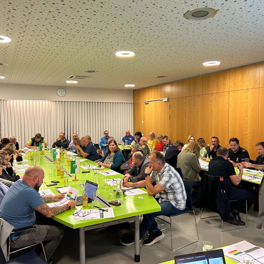 Eine große Gruppe von Menschen sitzt in einem Besprechungsraum um einen grünen Tisch und führt eine Diskussion. Der Raum hat Holzwände, eine weiße Decke mit Lichtern und eine Uhr. Einige Personen nutzen Laptops, während andere Papiere halten.