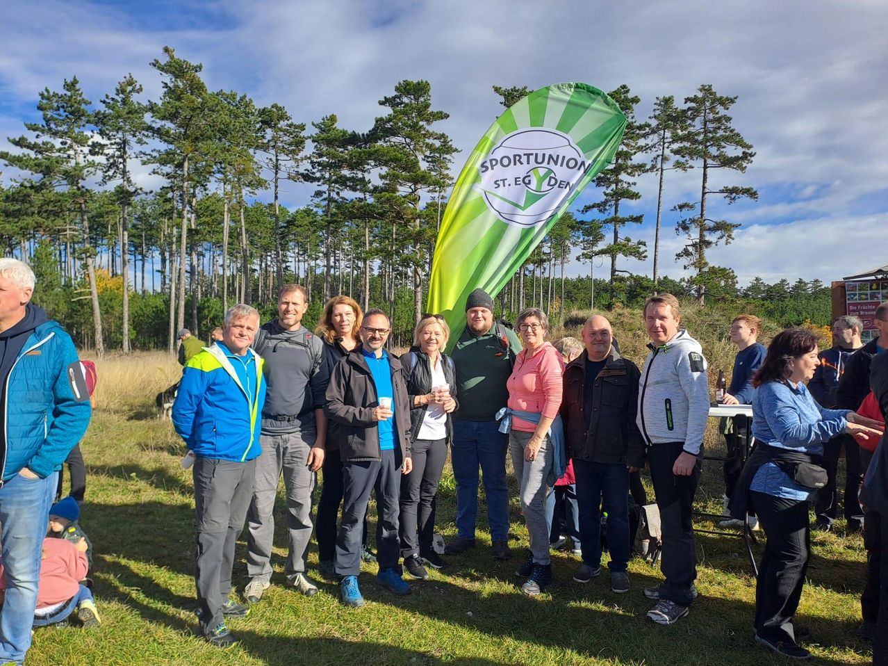 Eine Gruppe von Menschen steht zusammen auf einem Feld mit einem Banner, das Sportunion ST. EG. DEN liest. Sie sind für Outdoor-Aktivitäten gekleidet und lächeln für die Kamera.