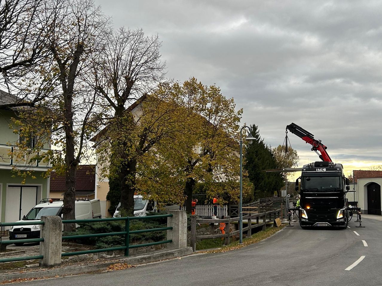 Ein Lkw mit einem Kran steht am Straßenrand. Es gibt Arbeiter und geparkte Fahrzeuge in der Umgebung. Bäume und ein Haus sind im Hintergrund.