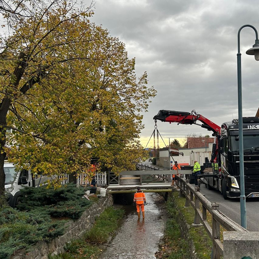 Ein Kran hebt einen Brückenabschnitt über einen Fluss. Arbeiter in orangefarbenen Westen stehen bereit. Im Hintergrund sind Bäume und ein bewölkter Himmel zu sehen.