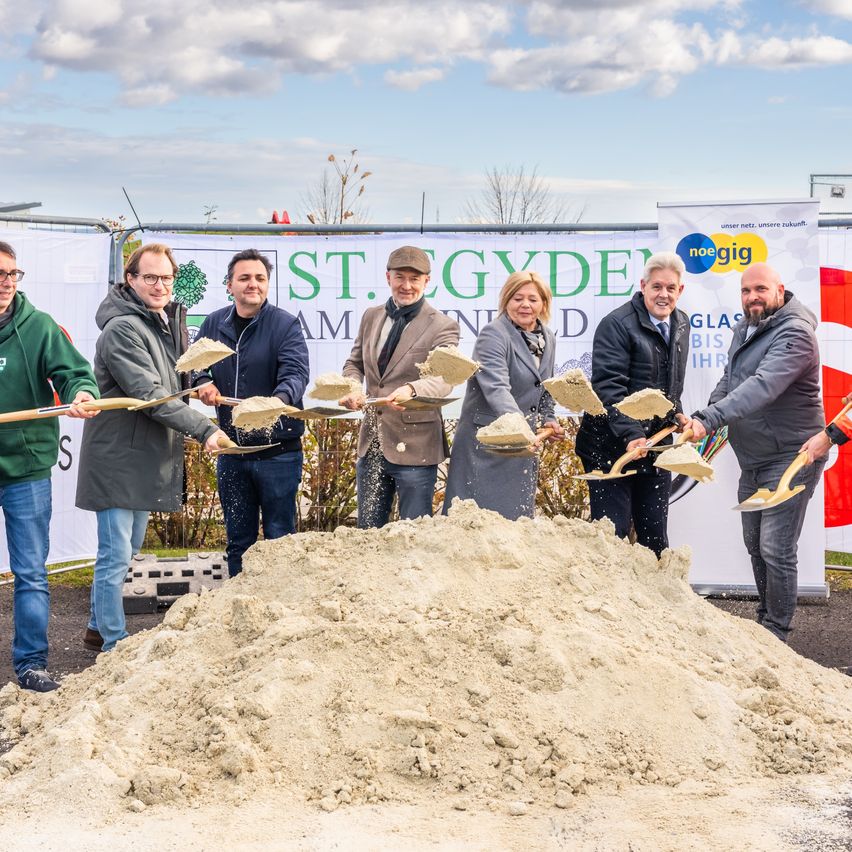 Eine Gruppe von Menschen schaufelt Erde an einer Baustelle. Dahinter befindet sich ein Banner mit dem Text 'St. Gydde'. Rechts sind eine Fahne und ein Banner angebracht.