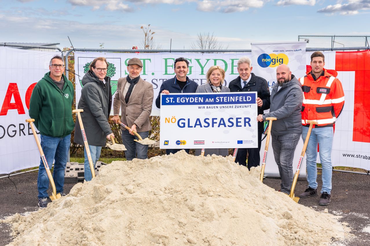 Eine Gruppe von Menschen in Mänteln steht um einen Haufen Sand herum und hält Schaufeln. Ein Schild zeigt 'St. Egyden am Steinfeld' und 'Glasfaser'. Sie befinden sich wahrscheinlich bei einer Grundsteinlegungszeremonie.