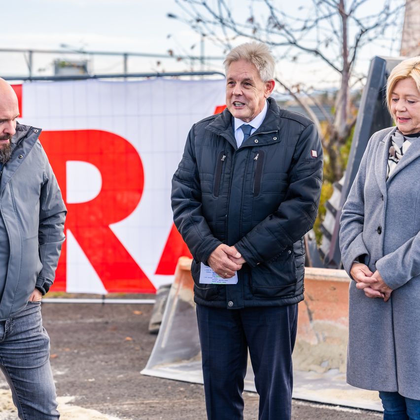 Drei Personen stehen vor einem Banner mit dem Buchstaben 'R'. Der Mann in der Mitte hält ein Stück Papier und trägt eine Krawatte. Hinter ihnen steht ein großer Bagger.
