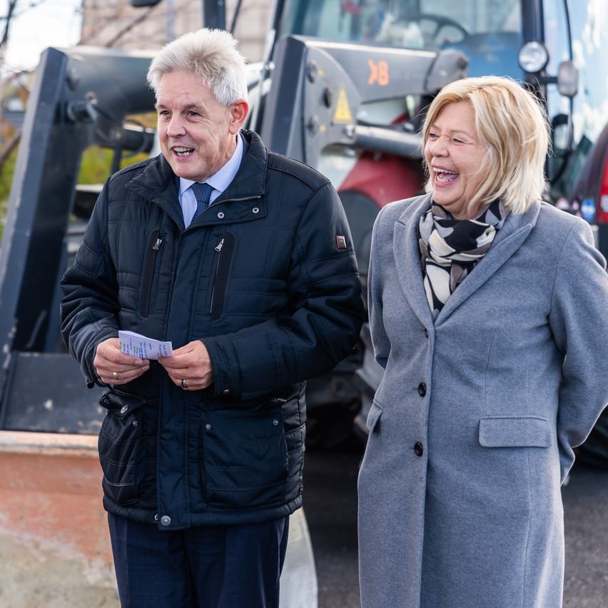 Ein Mann und eine Frau stehen neben einem Fahrzeug und lächeln. Der Mann hält ein Stück Papier in der Hand und trägt einen Anzug, während die Frau einen grauen Mantel mit einem Schal trägt.