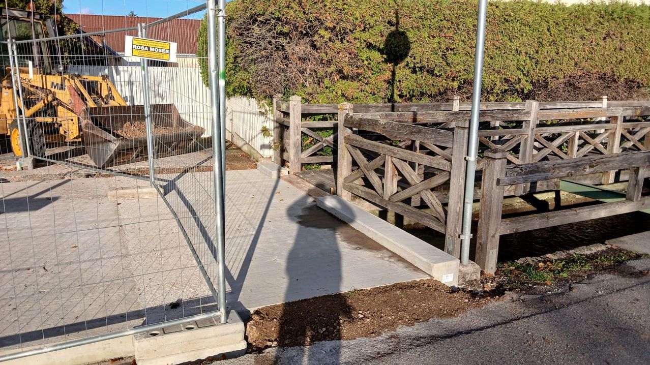 Eine Baustelle mit einer neuen Betonrampe, die zu einem hölzernen Tor führt. Ein Schatten einer Person wird auf den Boden geworfen. In der Nähe sind ein Metallzaun und ein Schild mit der Aufschrift 'Moser' zu sehen.