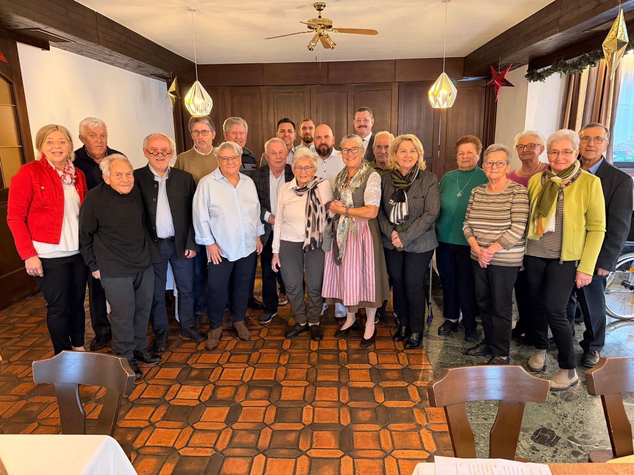 Eine Gruppe von Menschen in Winterkleidung, die in einem Raum mit Holzboden und Deckenventilator stehen. Einige halten Händchen.