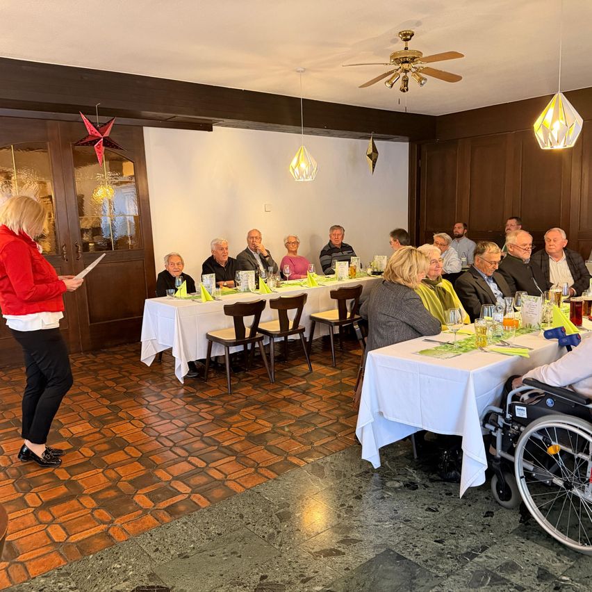 Eine Gruppe von Menschen sitzt an Tischen in einem Restaurant. Eine Frau steht davor und hält ein Blatt Papier. Der Raum hat Holzwände und eine Deckenventilator.