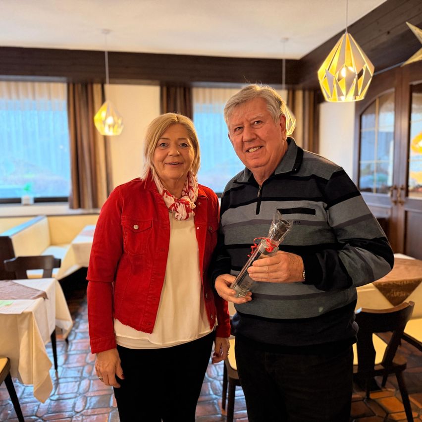Ein Mann und eine Frau stehen lächelnd zusammen in einem Restaurant. Der Mann hält ein Geschenk in einer Plastikverpackung. Hinter ihnen sind Tische und Stühle aufgestellt, und ein Fenster mit Vorhängen ist sichtbar.