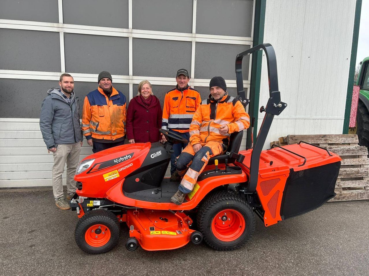 A group of people in orange safety suits stand next to an orange Kubota lawn mower in front of a garage.