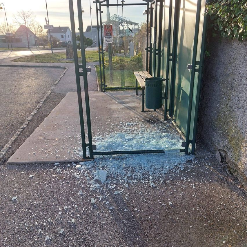A shattered bus stop with a green metal frame and a wooden bench is shown, with glass pieces scattered on the ground and a trash can nearby.
