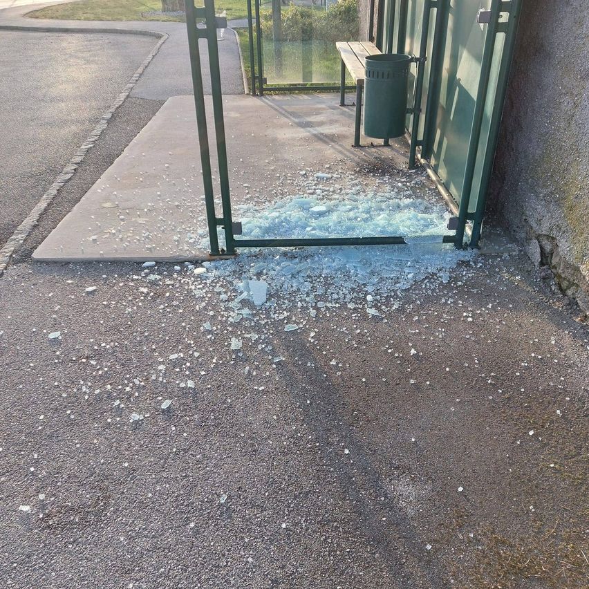 A bus stop shelter with a trash can has shattered glass on the ground. The glass is scattered around the area.