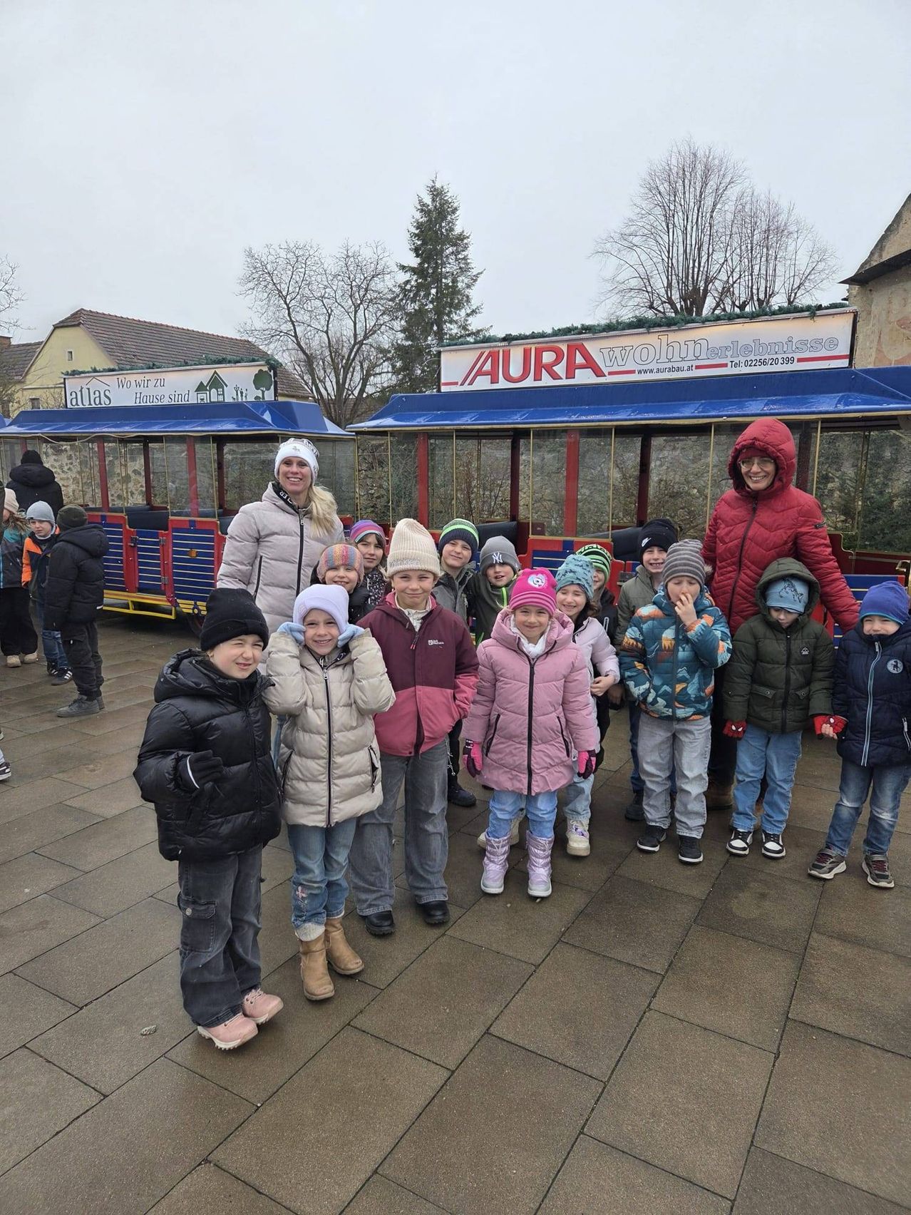A group of children and an adult woman stand in front of a colorful train, smiling for a photo. Behind them, a building with a sign reading AURA Wohnerlebnisse.