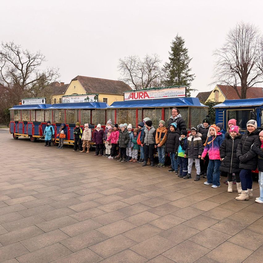 A group of children stands in line next to a train. They wear winter clothes. Behind them are buildings and trees.