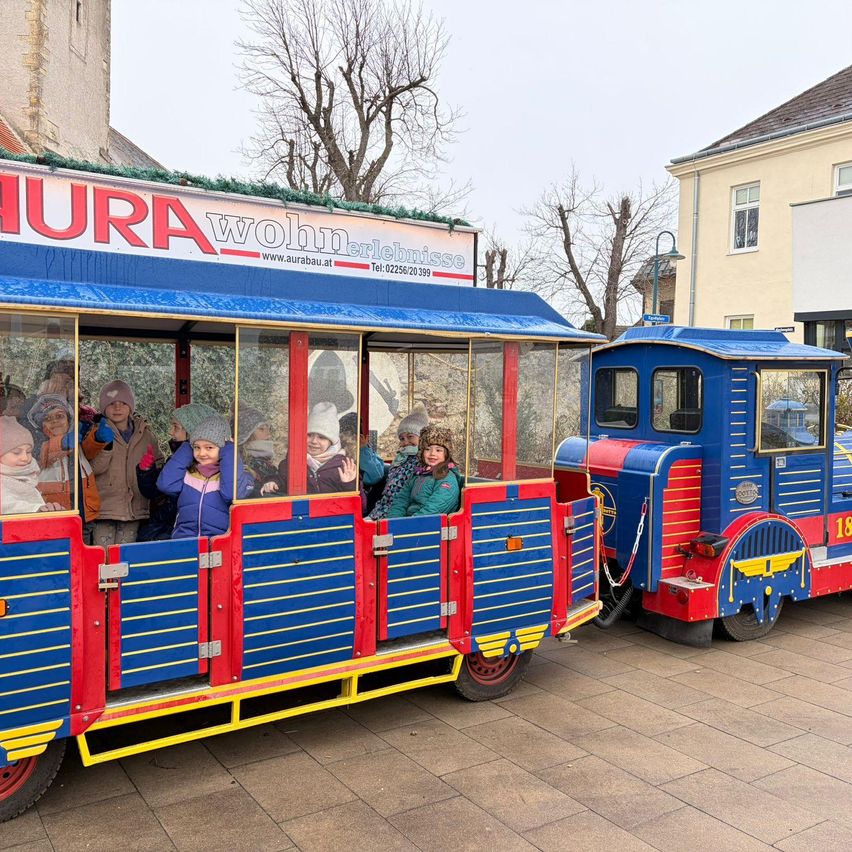 Children in winter clothing ride a colorful train with the number 18, parked in front of a building.