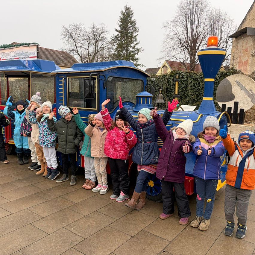 A group of children stands in front of a blue train, smiling and making gestures with their hands.