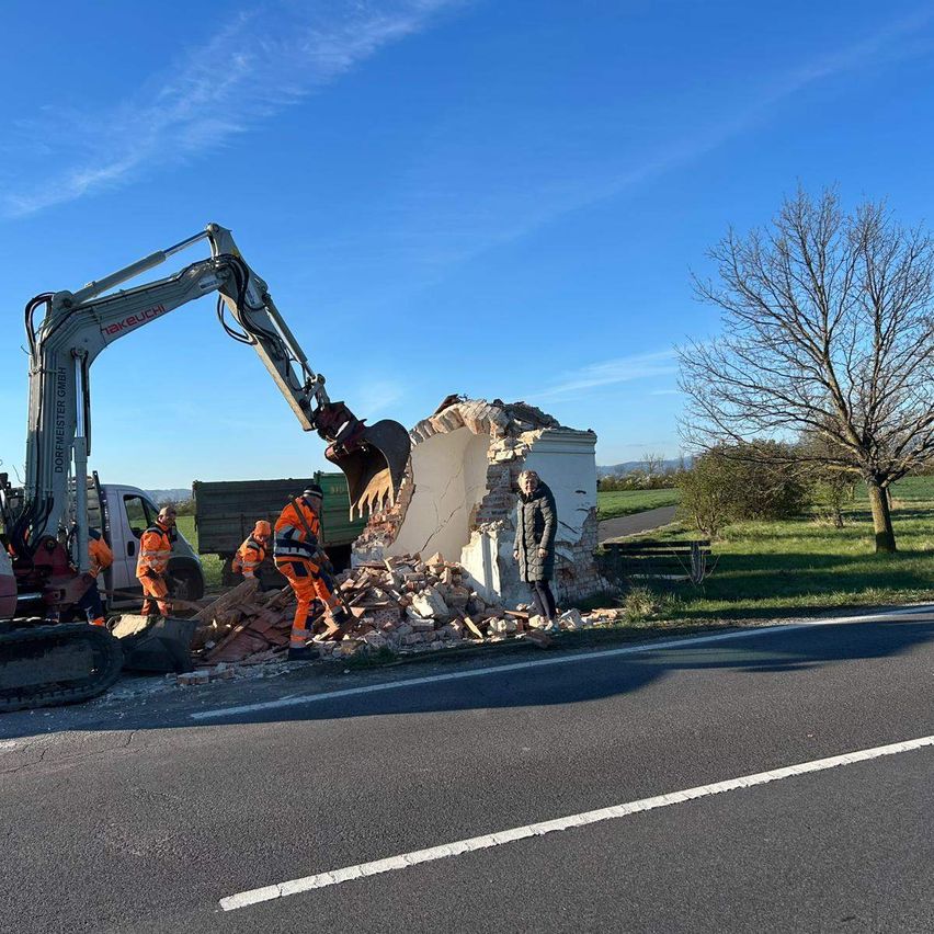 A road crew is demolishing a small structure by the side of a road using a large excavator.