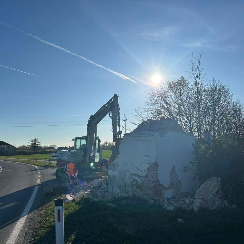A construction worker uses an excavator to demolish a damaged structure on the side of a road. The sun is bright and clear in the sky.