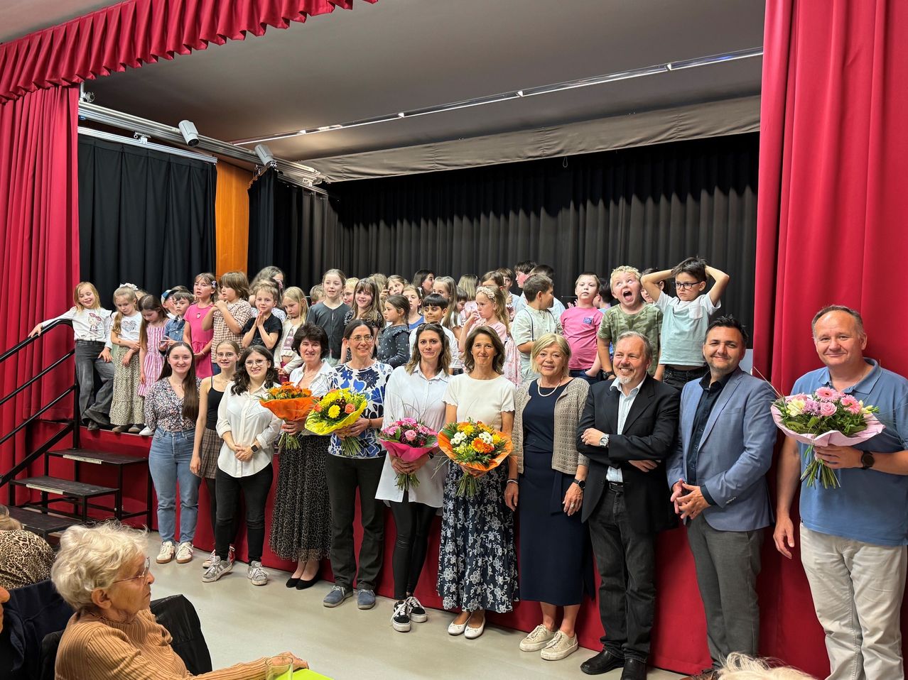 A group of people, including children and adults, stand on a stage holding flowers, likely celebrating an event with a red curtain backdrop.