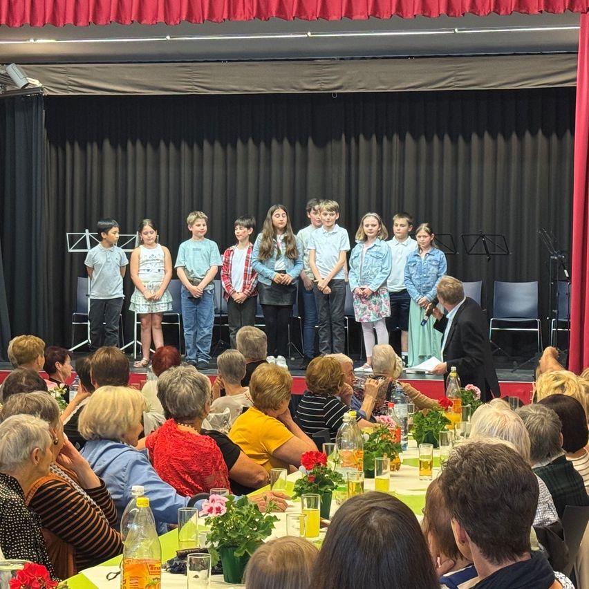 A group of children stand on a stage with a man behind a podium. The audience consists of older people sitting in chairs. The stage has black curtains, and there are flowers and drinks on the table in front.