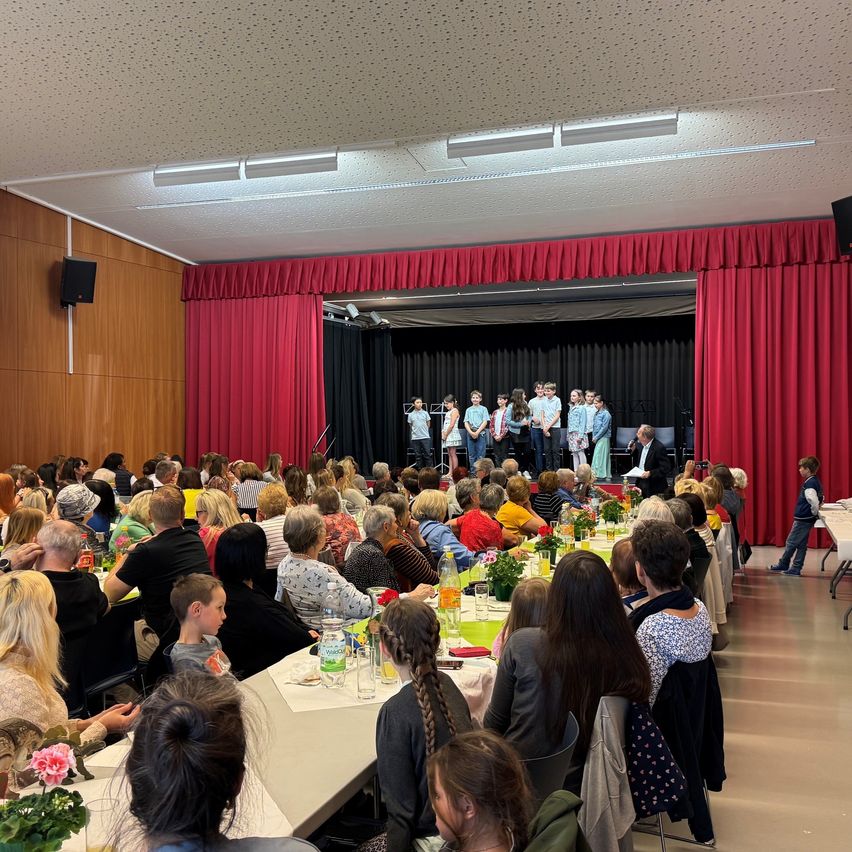 A room with a stage where a group of children and adults are performing. The audience sits at long tables with flowers and bottles. The stage is decorated with red curtains.