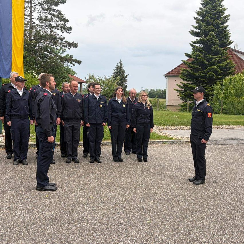 A group of uniformed individuals, possibly firefighters, are standing in a row in an open area. In front of them is a man with a serious expression. Behind them, there are some trees and a house.