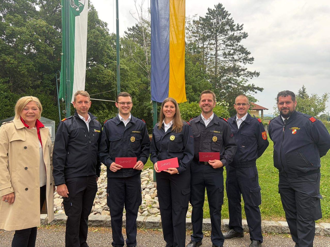 Five individuals, all in uniform, stand in a line holding certificates with the word 'DANKE'. They smile and seem to be posing for a photo. Behind them are two flags and some trees.