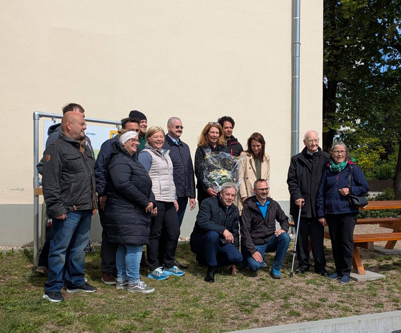 Eine Gruppe von Menschen posiert für ein Foto vor einem Gebäude. Ein Mann hält einen Blumenstrauß. Sie sind alle warm gekleidet, einige tragen Brillen und Mützen. Dahinter sind eine Bank und ein Baum zu sehen.