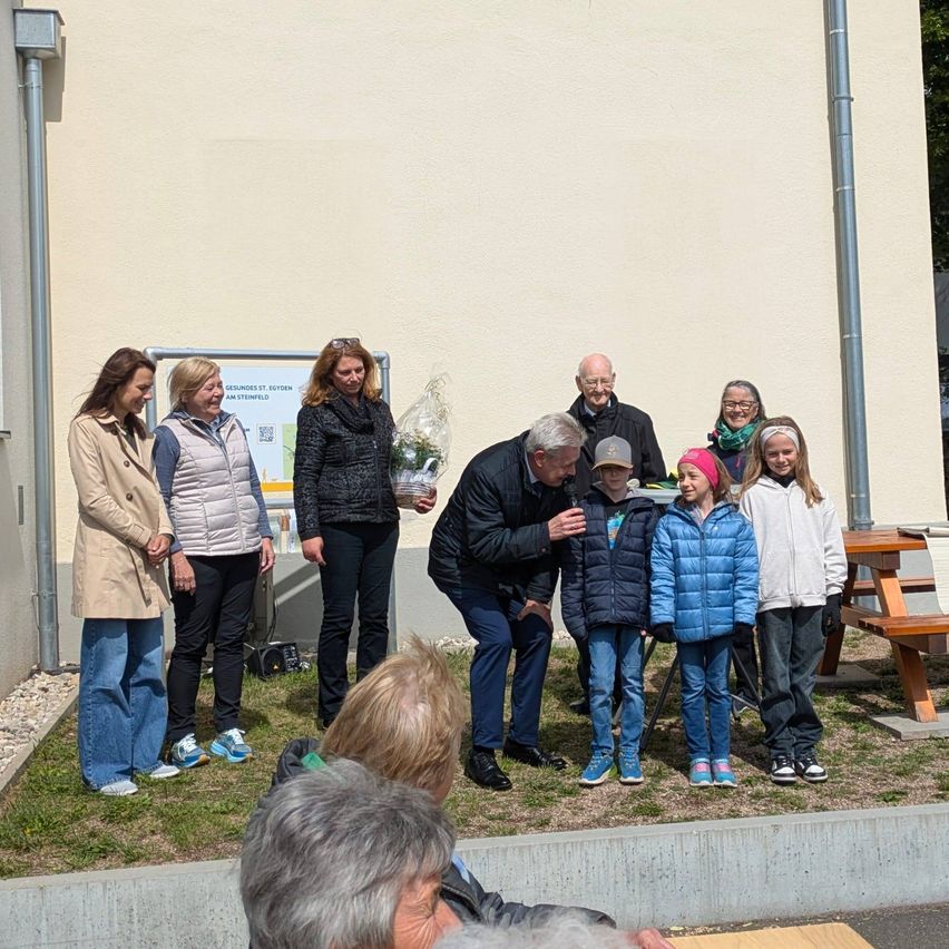 Mehrere Personen stehen auf einem Rasenbereich vor einem Gebäude. Dazu gehören ein älterer Mann, eine Frau mit einem Blumenstrauß und mehrere Kinder. Im Hintergrund ist eine Bank zu sehen.