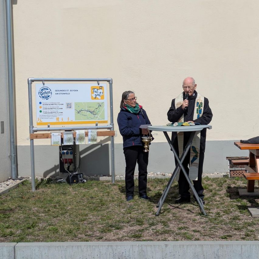 Ein Mann und eine Frau stehen neben einem Tisch mit Mikrofon und Schild, möglicherweise für eine Zeremonie oder Rede. Dahinter befindet sich ein Gebäude mit einer weißen Wand.