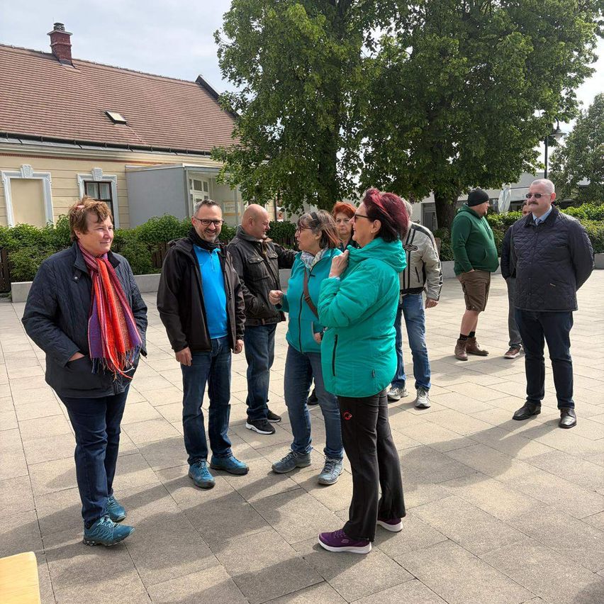 Eine Gruppe von Menschen steht im Freien und lächelt. Einige tragen eine Brille und eine Frau trägt einen Schal. Bäume und ein Gebäude sind hinter ihnen.