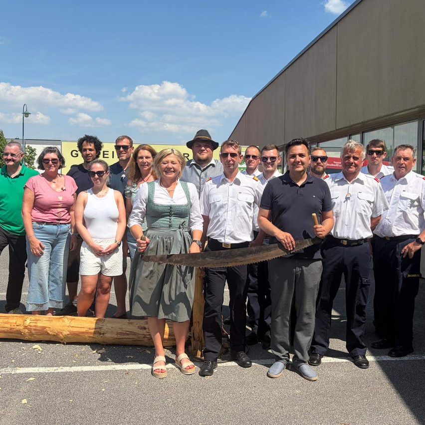 A group of people stand in a line, holding a large saw, and posing for a photo in front of a building.