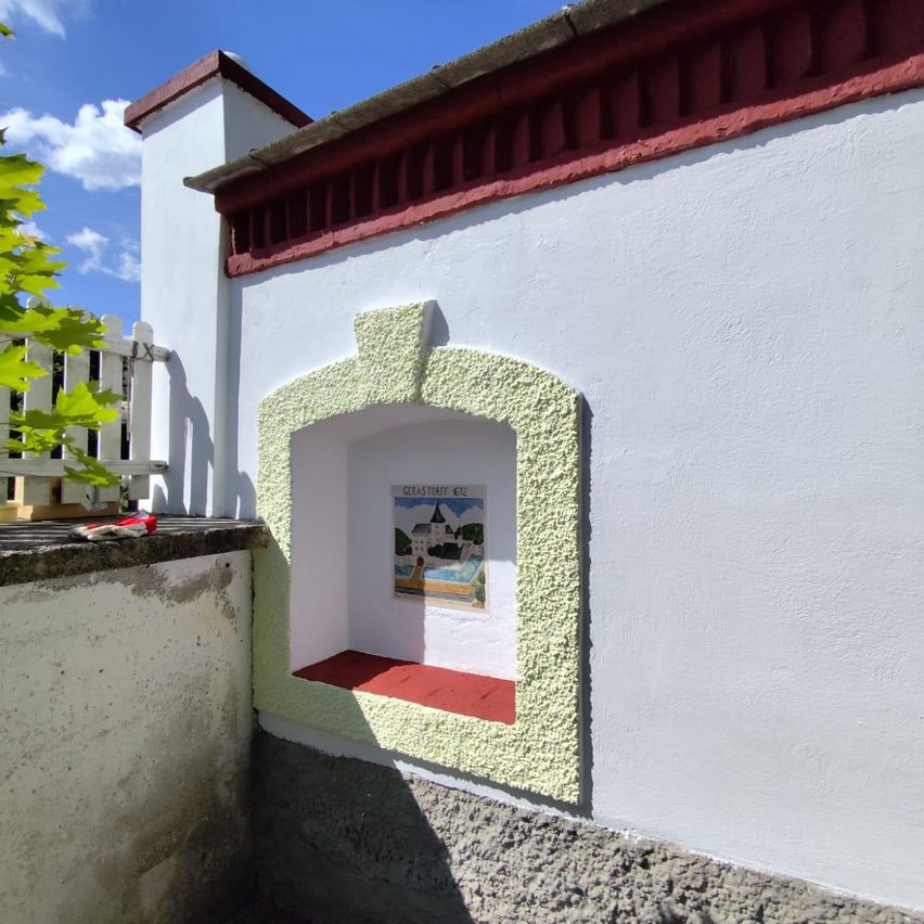 A white building with red trim has a small window with a red floor, showcasing a painting of a castle and a pool.