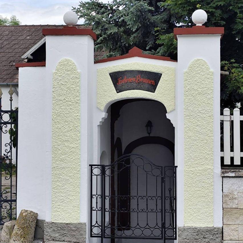 A white building with a red roof and arched doorway. The archway is labeled 'Fahrenheit Brunnen'. A black metal gate is in front of the doorway.