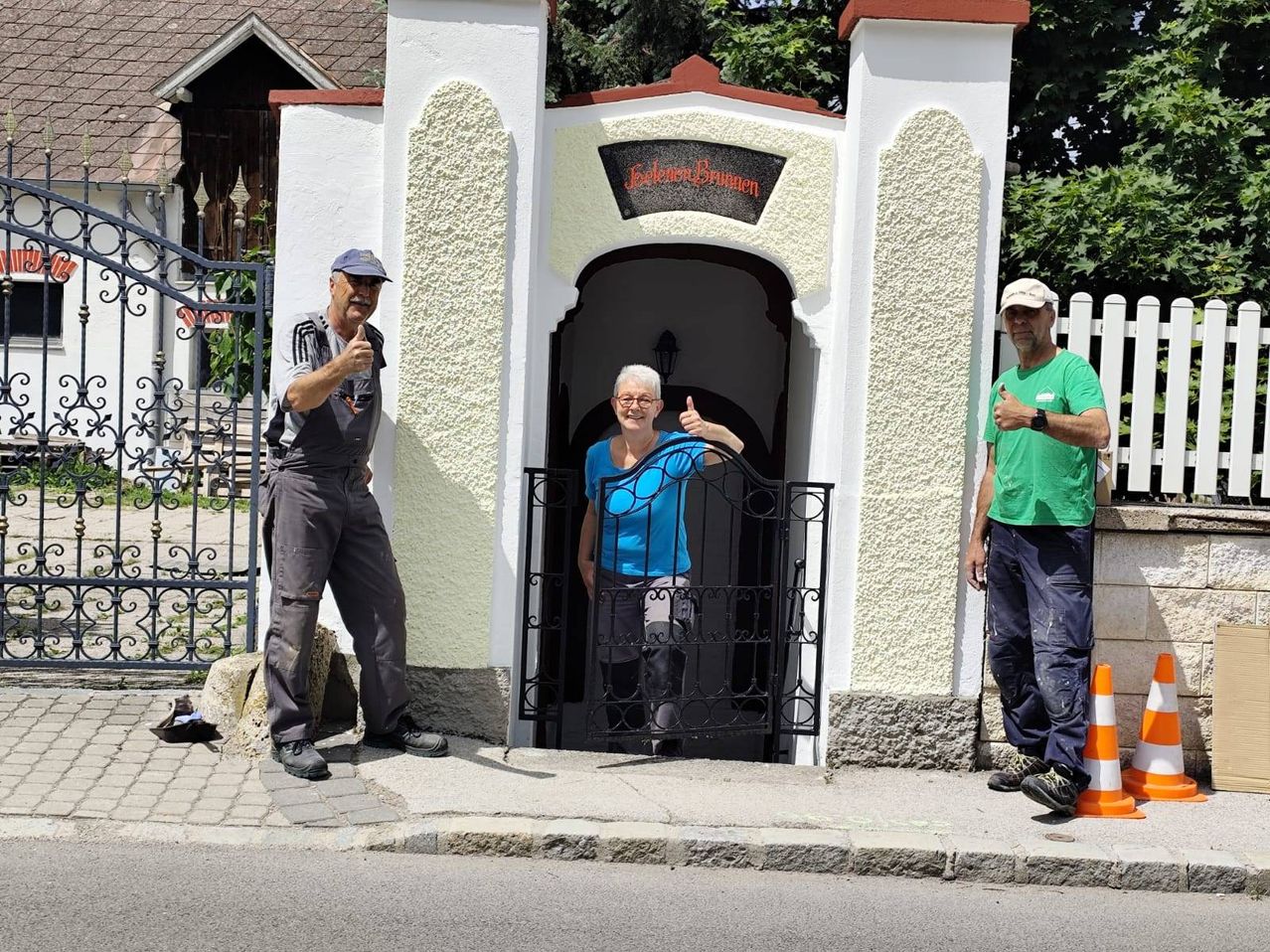 Three people stand in front of a building with an arched entrance. The woman in blue stands in the entrance. Both men wear thumbs up.