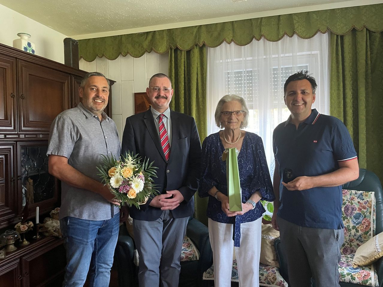 Vier Erwachsene, drei Männer und eine ältere Frau, stehen in einem Raum mit einer Blumenarrangement und einer grünen Geschenktüte, lächeln für ein Foto.