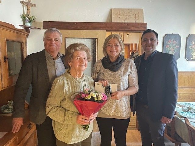 Four people stand together in a room. A woman holds a bouquet of flowers. The woman beside her holds a certificate. Behind them, a cross and picture frame are on the wall.