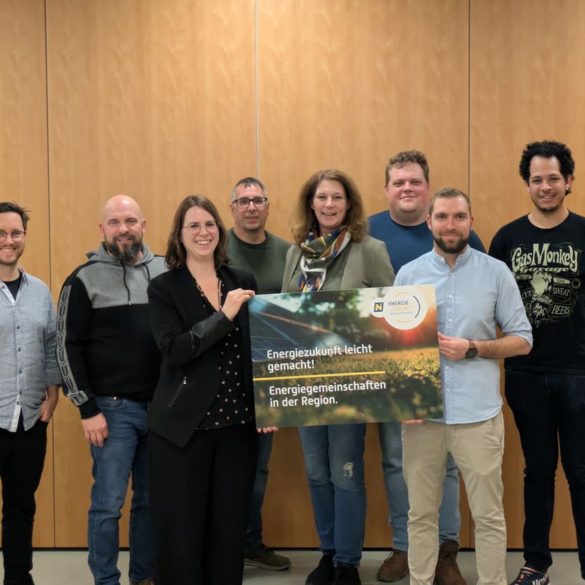 Group of people standing and smiling, holding a sign that reads 'Energiezukunft leicht gemacht! Energiegemeinschaften in der Region.' They are in front of a wooden wall.