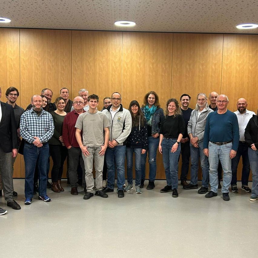 Eine Gruppe von Menschen steht vor einer Holzwand. Sie sind alle lässig gekleidet. Der Mann in der Mitte trägt eine Brille und eine Uhr. Die Frau neben ihm trägt einen Schal.