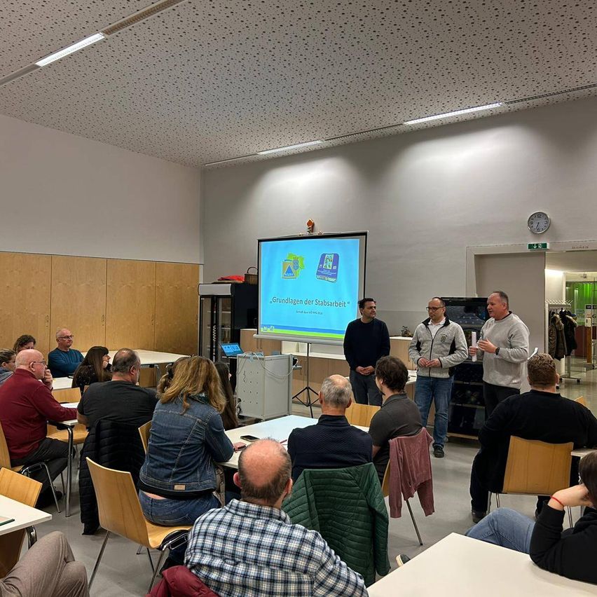 Eine Gruppe von Menschen befindet sich in einem Besprechungsraum mit einer Projektionsleinwand, auf der eine Präsentation läuft. Einige Personen stehen und sprechen in der Nähe der Leinwand. Der Raum ist gut beleuchtet, an der Wand hängt eine Uhr.