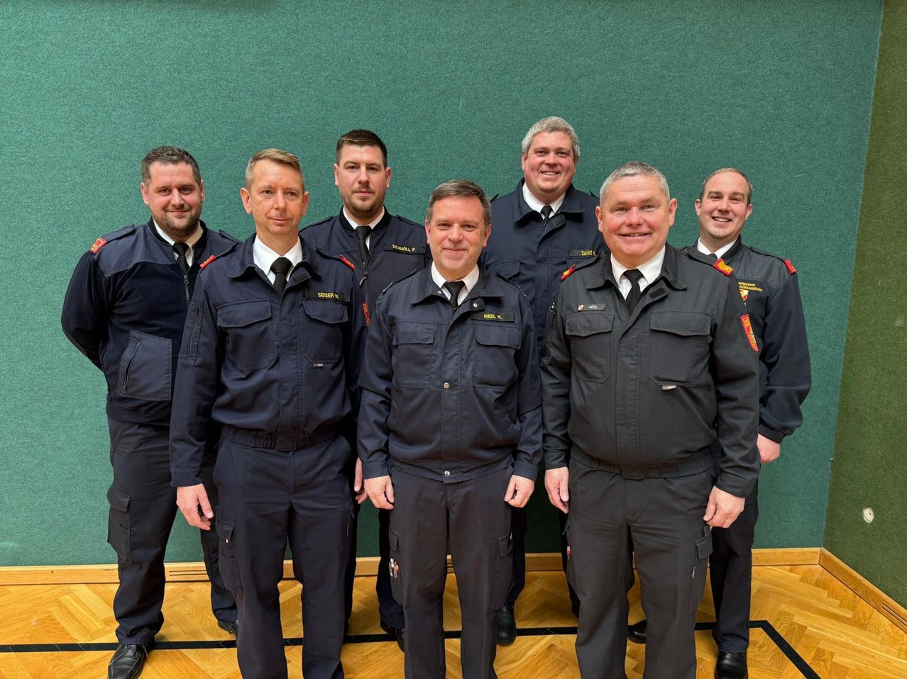 Eine Gruppe uniformierter Feuerwehrleute posiert für ein Foto vor einem grünen Hintergrund, alle in schwarzen Uniformen und Krawatten.