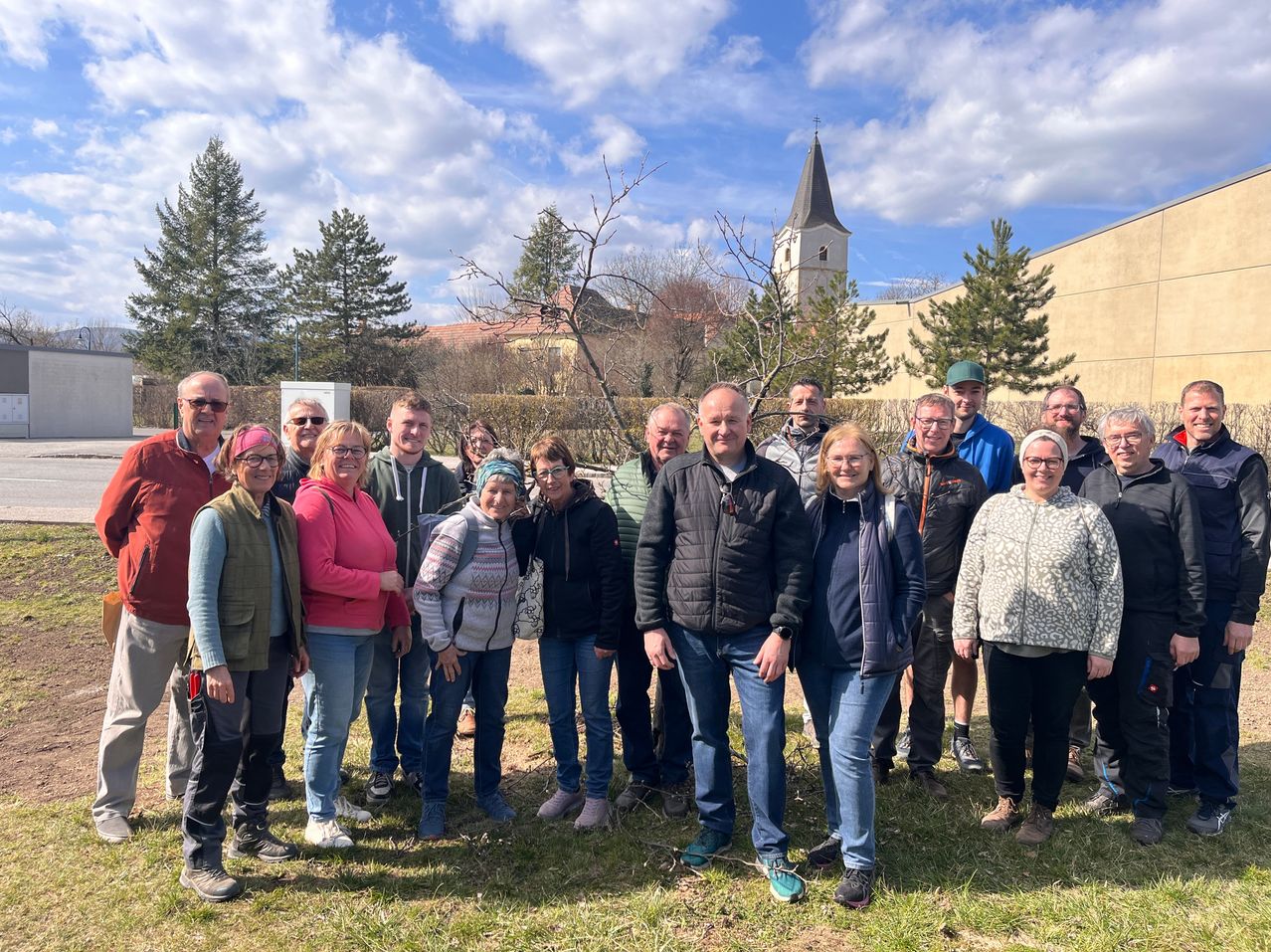 Eine Gruppe von Menschen in lässiger Kleidung steht vor einem Gebäude mit einem Turm unter einem bewölkten Himmel. Im Hintergrund sind Bäume sichtbar.